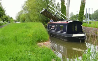 Narrowboat named 'Jam' passes under a lift bridge on the Oxford Canal, surrounded by a verdantly grassy towpath