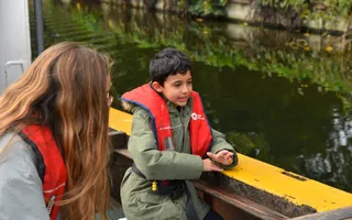 A young boy with dark hair in a green coat and red life jacket leans on the side of a yellow boat, looking out at the canal.