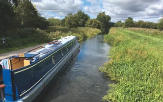 Narrowboat moves along a rural canal with vegetation sticking into the canal and a grassy towpath.