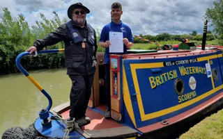 Two men stand by the tiller of a narrowboat, one holding a certificate