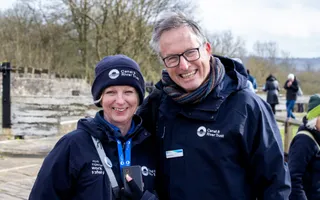 Two people wearing dark blue coats, person on the left wearing wool hat, both smiling to camera