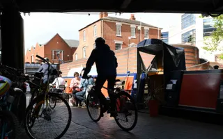 Person on bike passes moored narrowboats under a bridge.