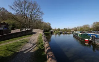 To the left, a towpath leads up and over a bridge which disappears off, whilst a wide blue canal with boats runs parallel.