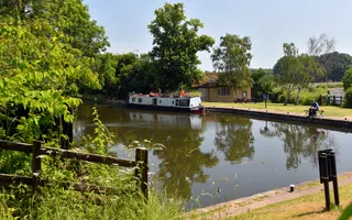 A warm sunny day, a boat moored on the right of a canal with a man in a t-shirts fishing close by. The canal is surrounded by grass and trees.
