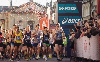 A group of runners race past cheering spectators against Oxford's scenic buildings.