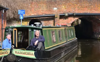 Two people sit smiling at the front of their moored narrowboat as they fill up with water.