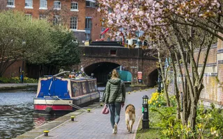 Woman walks dog down the towpath towards a bridge, passing a moored boat.