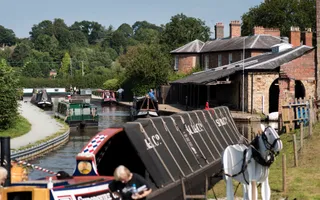 A traditional working boat with sloped canvas sides is moored along a grassy bank with a wooden horse.