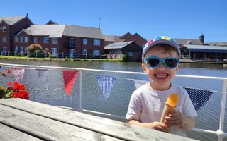 Smiling boy wearing sunglasses and cap sitting next to water holding an ice lolly
