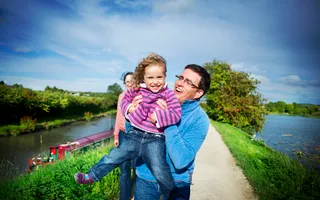 Father holding up daughter on a towpath with water on each side on a sunny day.
