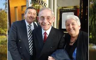 An elderly man in a suit and glasses smiles at the camera, with a younger man in a suit to his left and a woman with white hair to his right.