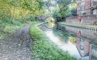 a canal leading towards a bridge with brick building on the right and reflections in the water. Muddy towpath running parallel on the left