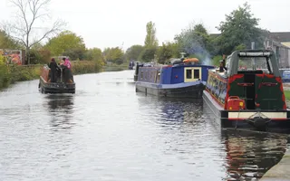 Narrowboat passes moored boats with smoke blowing from chimneys, surrounded by trees.