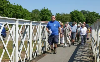 Volunteer leads a group of visitors across an old iron swing bridge on a sunny day