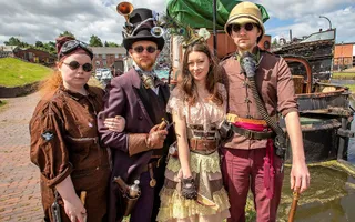 Four people stand arm in arm dressed in steampunk outfits in front of old work boats.