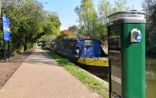 A green electric pillar for charging eco boats is positioned to the right of the picture, behind it moored boats are seen stretching back with a gravel towpath running parallel to the left.