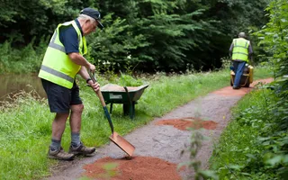 A man with a shovel stands on towpath next to a wheelbarrow surrounded by greenery