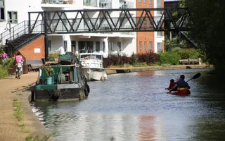 A man and child kayak pass moored boats, flats, and a cyclist on the towpath, under a footbridge, on a sunny day.