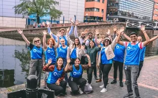 A group of people in office wear with 'Canal & River Trust' branded running vests over the top stand by the canal in the centre of Birmingham smiling with their hands raised in celebration.