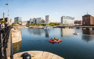 A large expanse of water, tall buildings in the horizon, and people paddling in a red double canoe in the foreground.