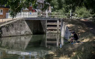 A man sits on the canal bank and fishes in front of a lock gate.