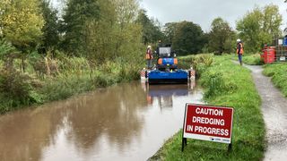 Wide shot of a canal and towpath with dredging boat in the background and caution sign in the foreground