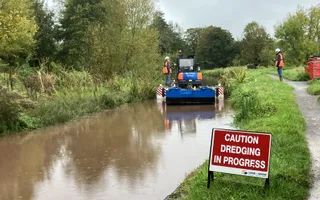 Wide shot of a canal and towpath with dredging boat in the background and caution sign in the foreground