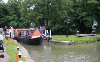 A red trip narrow boat leaves a lock at a canal junction at Cosgrove with onlookers watching and volunteers helping..