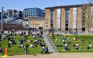 Groups in twos or more sit on grassy steps enjoying food in the sun against the backdrop of London's King's Cross.