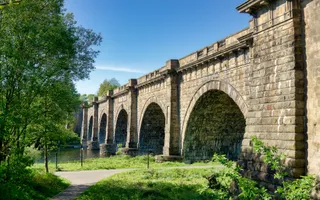The sun shines on footpath passing a stone aqueduct that carries the canal over a wide river.