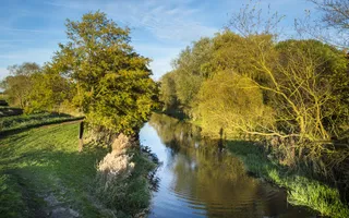 Sun sets over a rural canal, lined by trees and fields.