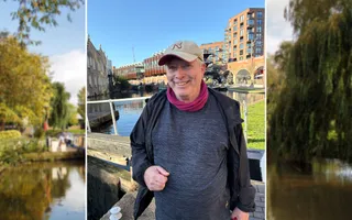 A man in a baseball cap, navy top and black coat smiles at the camera in front of a lock gate on a canal.
