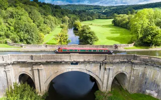 Aerial view of a narrowboat crossing a stone aqueduct over a river and fields in the shining sun.