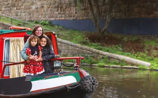 A woman holds two children close as they stand on the front of a canal boat, smiling.