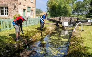 Two men in blue hi-vis and red lifejackets stand on the grass verge, leaning in with long rakes to scoop out weeds from the water.