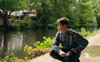A young boy with dark brown hair in a grey hoodie crouches down on the towpath and looks out towards the canal.