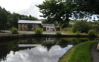 Woman on bike cycles round a corner of a towpath where the surrounding trees and brick building reflect in the canal