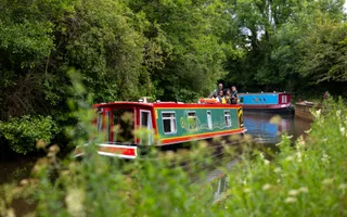 A red and green narrowboat cruises along calm, rural waters with thick greenery covering the towpath.