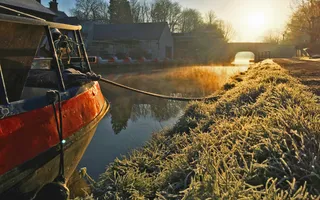 On the left, the red bow of a boat is moored alongside frosty grass which stretches back towards a bridge, with the sunset casting an orange glow