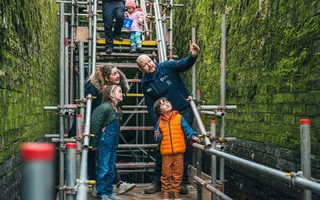 Family stand on scaffolding in a drained lock as a Canal & River Trust volunteer explains the works