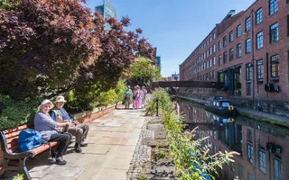 Two elderly people sitting on a bench beside a canal on a sunny day