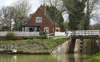 Brick building sits next to the canal below a lock, with people walking by.