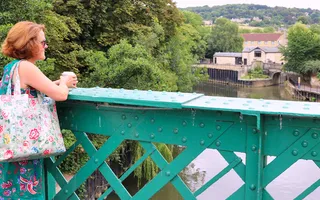 Woman in flowery dress with bag looks over a metal bridge towards the canal.