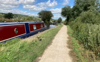 A flat gravel towpath with a grass bank runs alongside the canal. A red and blue narrowboat is moored up on the water.
