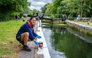 A dark-haired woman with a blue jumper and red lifejacket crouches down to paint the lock edges with white paint