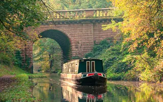 A narrowboat passes under a high, arched brick bridge in dappled autumnal sunlight.