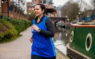 A smiling woman in a branded running vest jogs along the towpath, passing a moored narrowboat.