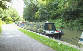 Line of moored boats on the towpath, surrounded by trees and greenery on a sunny day.