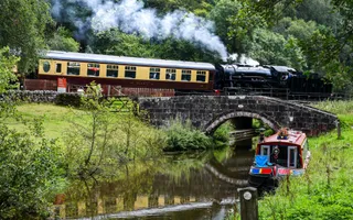 A steam train crosses the canal where a narrowboat is moored.