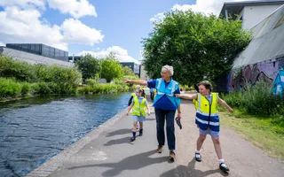 A woman in a blue hi-vis walks next to school children in blue dresses and yellow hi-vis, all are pointing across the canal to something on the other side.